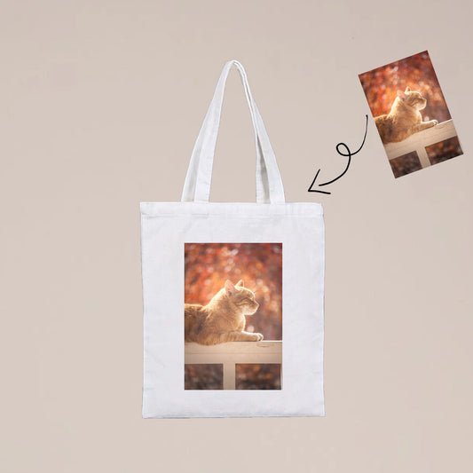 A photo of a custom photo tote bag. The tote bag is white and has a photo of a cat on it. The cat is a brown tabby cat.